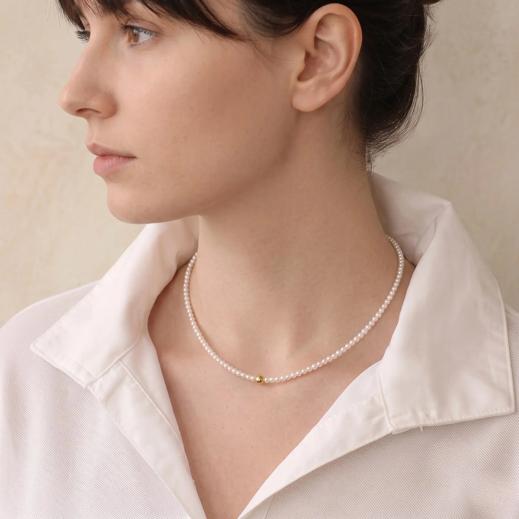 Side profile of a woman wearing a delicate freshwater pearl necklace, highlighting the luminous pearl strand and gold center detail.