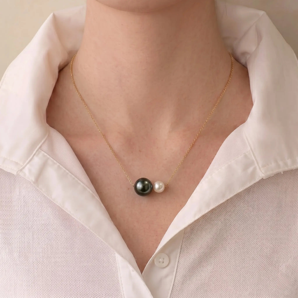 A close-up of a person wearing a delicate gold chain necklace with two pearl-like beads, one dark and one light, styled with a white collared shirt.