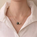 A close-up of a person wearing a delicate gold chain necklace with two pearl-like beads, one dark and one light, styled with a white collared shirt.