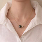 A close-up of a person wearing a delicate gold chain necklace with two pearl-like beads, one dark and one light, styled with a white collared shirt.