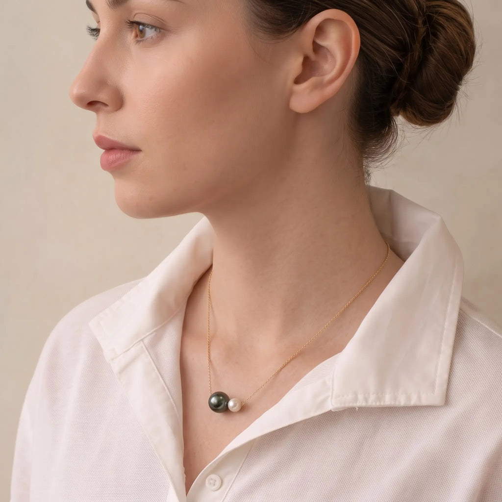 A close-up side profile of a woman wearing a delicate gold chain necklace with a pendant made of one dark black pearl and one small white pearl, paired with a white collared shirt.