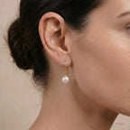 A close-up of a woman's ear wearing a delicate gold dangle earring with a single white pearl drop, against a soft beige background.