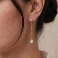 Side profile view of a woman wearing fluid 18k solid gold pearl drop earrings with an unbuttoned white linen shirt, demonstrating how the chain visually sculpts the jawline.