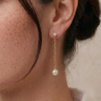 Side profile view of a woman wearing fluid 18k solid gold pearl drop earrings with an unbuttoned white linen shirt, demonstrating how the chain visually sculpts the jawline.