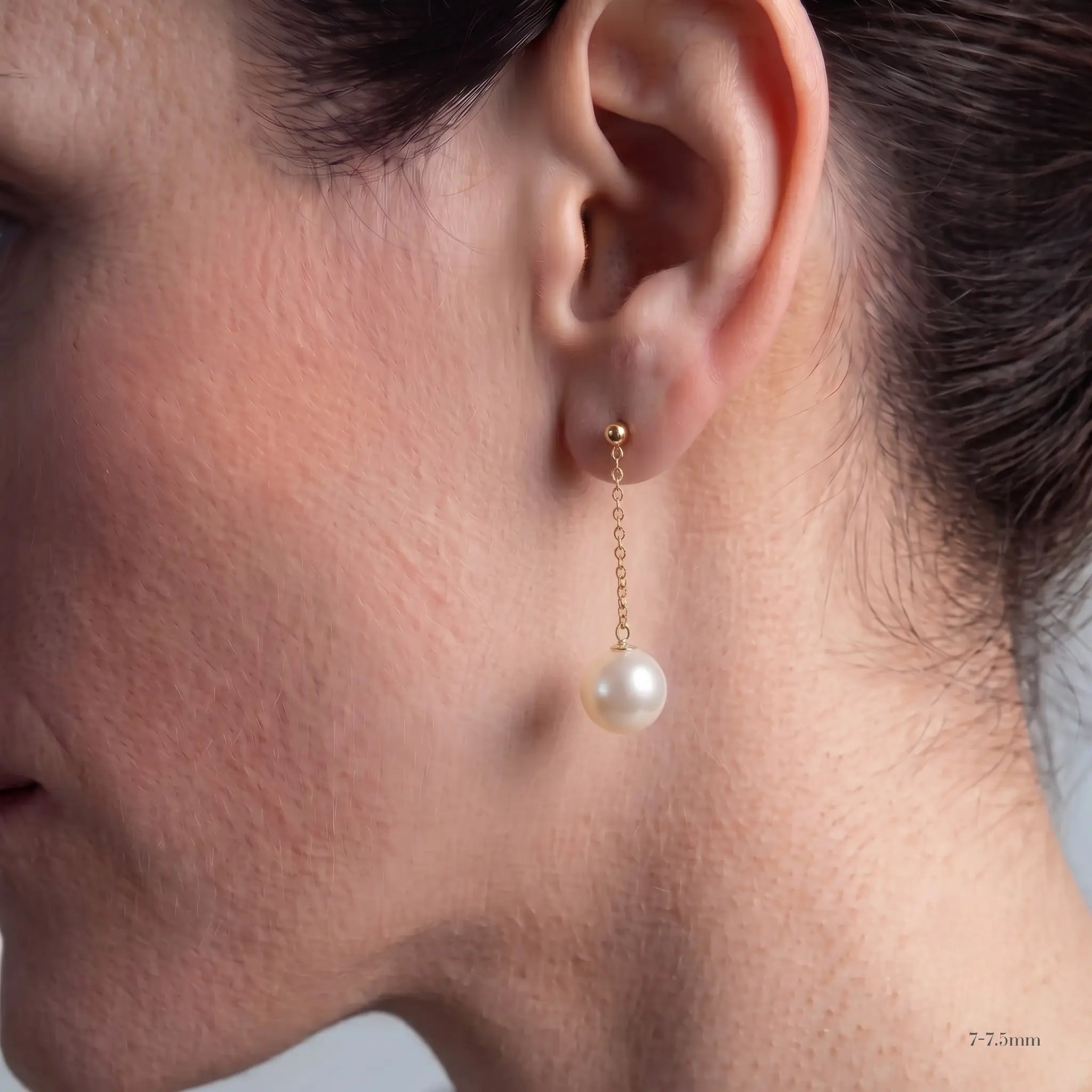 A close-up side profile of a woman wearing a single gold drop earring with a white pearl pendant. The image highlights the earring on the ear, with the model's skin, hair, and neck visible against a neutral light gray background.