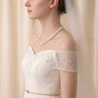 A woman in profile wearing a bridal outfit with an off-the-shoulder cream dress, sheer embroidered sleeves, a double-strand pearl necklace, and a long white veil against a soft cream curtain background.