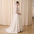A woman wearing a long ivory wedding dress with a matching veil stands in profile against a cream curtain backdrop in a bridal shop.