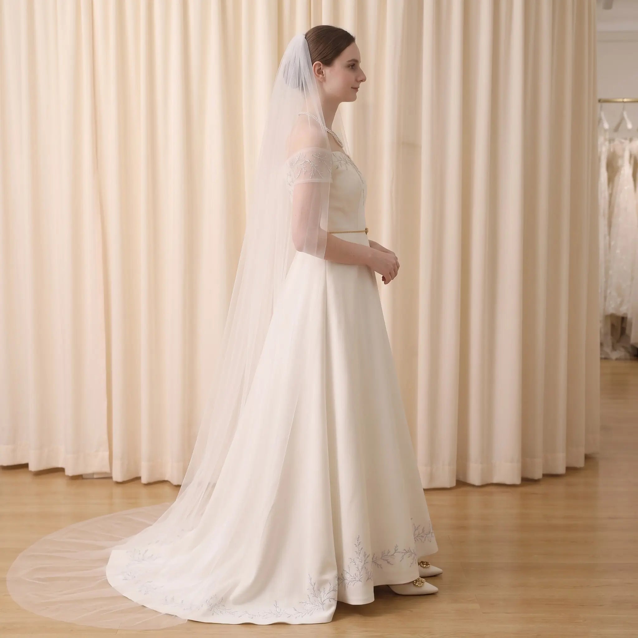 A woman wearing a long ivory wedding dress with a matching veil stands in profile against a cream curtain backdrop in a bridal shop.