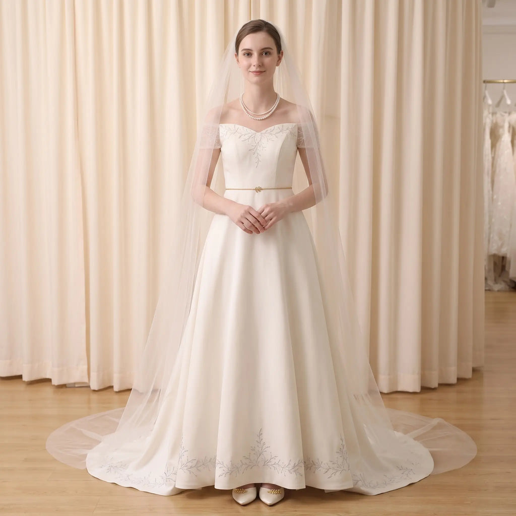 A woman stands in a bridal gown with a long sheer veil, photographed indoors in front of cream curtains. The dress is off-the-shoulder with a fitted bodice, a thin waistband, and a full skirt with embroidered detailing near the hem.