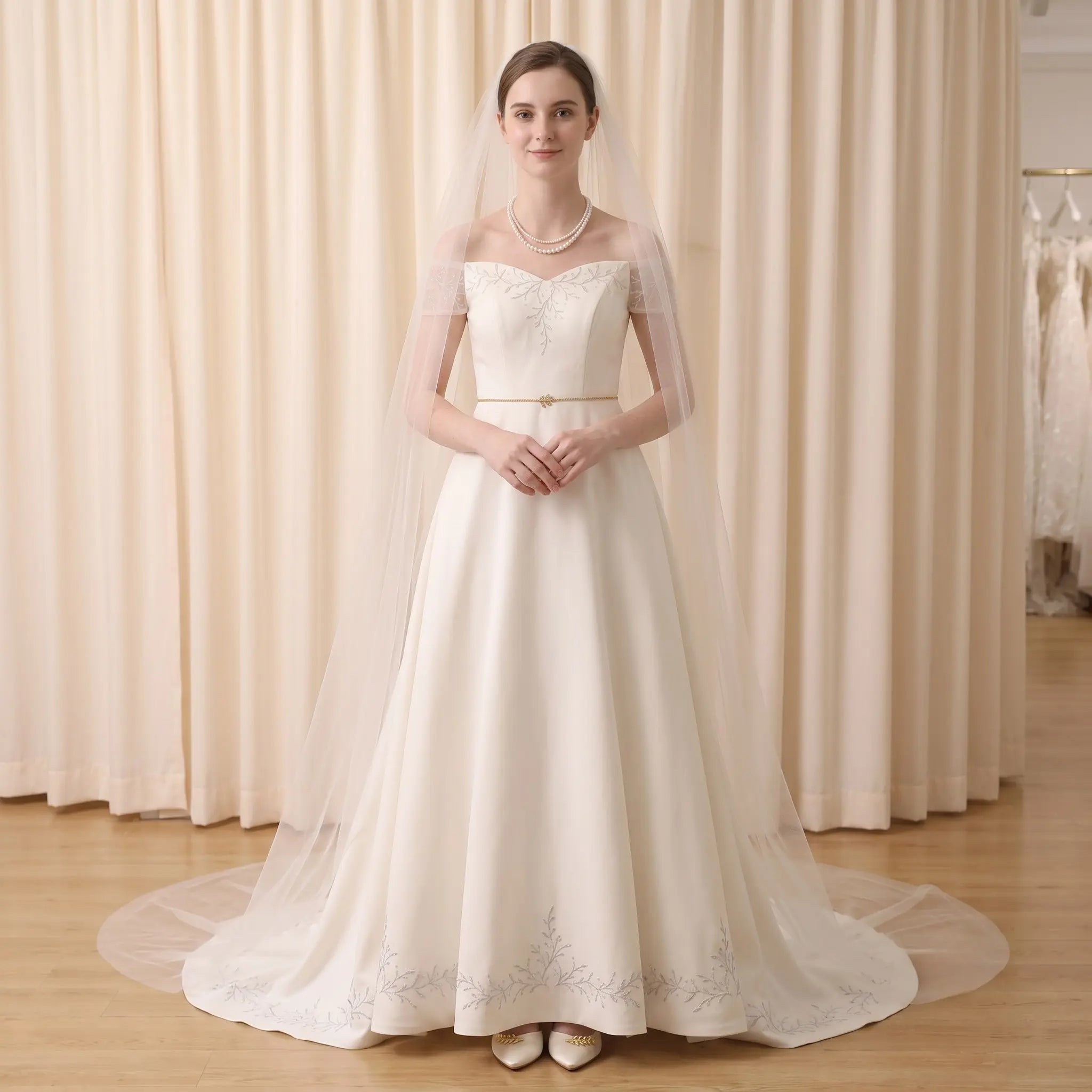 A woman stands in a bridal gown with a long sheer veil, photographed indoors in front of cream curtains. The dress is off-the-shoulder with a fitted bodice, a thin waistband, and a full skirt with embroidered detailing near the hem.