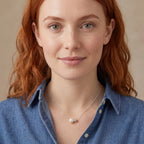 A close-up portrait of a woman with red hair wearing a delicate silver necklace with a single pearl and a small gold bead, paired with a blue denim shirt against a soft beige background.