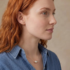 A close-up side profile of a woman with red hair wearing a delicate silver necklace with a white pearl pendant, paired with a blue denim shirt against a neutral beige background.