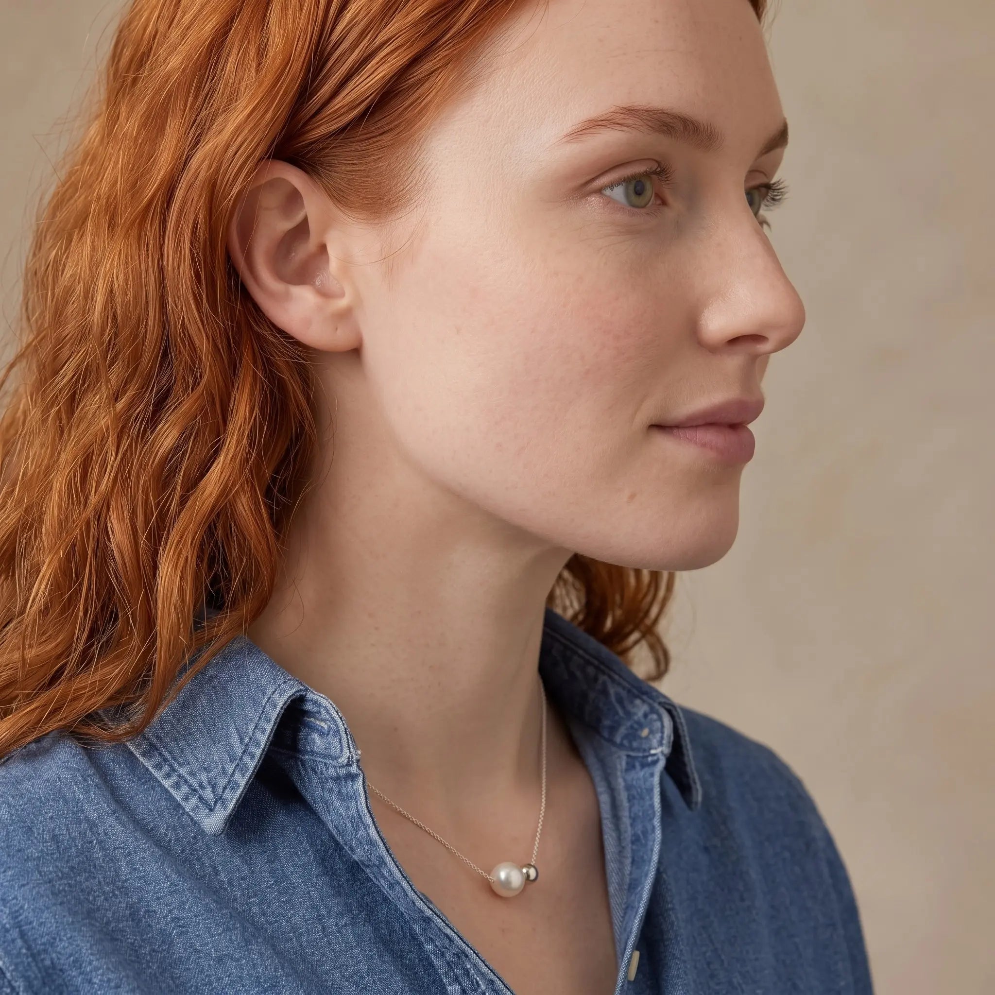 A close-up side profile of a woman with red hair wearing a delicate silver necklace with a white pearl pendant, paired with a blue denim shirt against a neutral beige background.